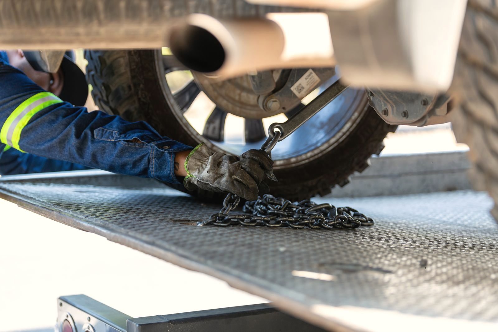 An operator kneeling to fasten securing chains under a vehicle on a flatbed deck